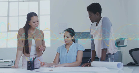 Pointing three women in office attire leaning on meeting table, reviewing rolled plans with pen cup....