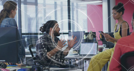 Gesturing bearded man with bun in plaid shirt leading meeting at open-plan office, with monitors. Collaboration, teamwork, discussion, devices, daylight, modern, coworkingの写真素材