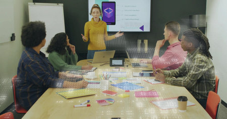Presenting woman in mustard sweater gesturing at conference table, with laptop and sticky notes....