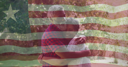 Embracing mature couple standing in sunlit park, man kissing woman in red plaid shirt, flag overlay. Duo, outdoors, hug, affection, warmth, garden, natureの写真素材