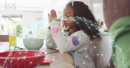 Praying girl in knit sweater closing eyes and folding hands at dining table, digital mesh overlay. Child, breakfast, bowl, orange, juice, family, lifestyleの写真素材