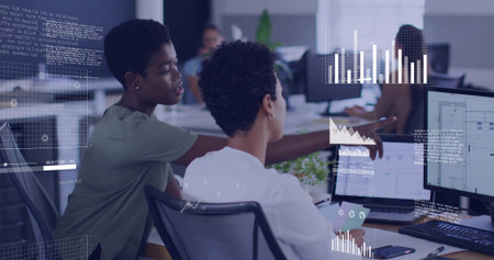 Pointing coworker in green top guiding colleague in white blouse at office, dual monitors overlays....