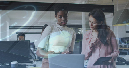 Pointing, woman in ribbed top and colleague in pink blouse reviewing laptop at office, with tablet. Collaboration, teamwork, professionals, workspace, monitors, digital, gridの写真素材