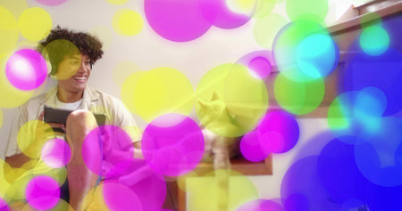 Sitting man in white tee smiling holding tablet on steps by window, cat resting, colorful bokeh. Bench, device, pet, daylight, interior, cushions, calmの写真素材