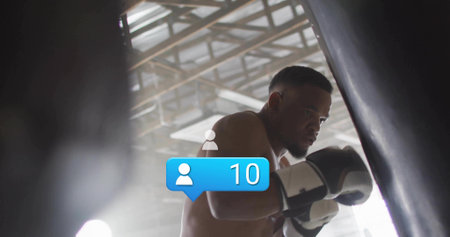 Punching shirtless boxer leaning into bag at boxing gym, wearing gloves and wraps. Athlete, focus, intensity, training, discipline, industrial, hazyの写真素材