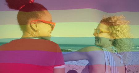 Talking women facing each other at beach, wearing shades, red tee, striped swimsuit. Shoreline,...