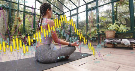 Sitting woman on yoga mat in greenhouse in sports top and smartwatch. Meditation, serenity, glasshouse, tropical, plants, mindfulness, fitnessの写真素材