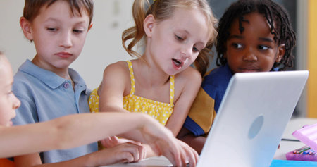 Pointing blonde girl in yellow dress reading at school table, with silver laptop, pink pencil case. Classroom, students, collaboration, group, device, screen, learningの写真素材