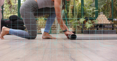 Kneeling grey-clad woman rolling up exercise mat on glass sunroom floor with bolster, plants, watch. Yoga, fitness, wellness, balance, focus, serenity, woodenの写真素材