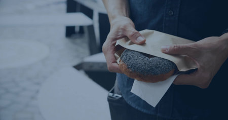 Holding mid-adult man in dark shirt pulling back paper wrapper at cafe plaza, with seeded pastry. Donut, outdoor, urban, unwrapping, pouch, closeup, calmの写真素材