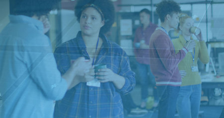 Listening mid-adult woman in blue plaid shirt holding mug at open-plan office, lanyards visible. Coworking, collaboration, conversation, teamwork, monitors, daylight, modernの写真素材
