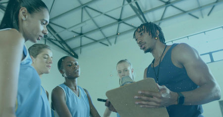 Talking coach in dark tank holding clipboard at gym with players in light-blue jerseys, copy space. Basketball, huddle, timeout, teamwork, leadership, strategy, courtの写真素材