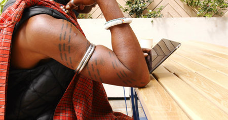 Leaning man tattooed arms with bracelets wearing plaid at wooden table in patio, with tablet cup. Courtyard, outdoor, alfresco, portrait, casual, relaxed, lifestyleの写真素材