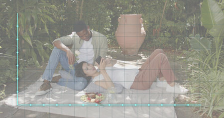 Lying woman holding phone, man leaning back wearing casual clothes on blanket in garden, fruit bowl. Couple, outdoors, secluded, lush, tropical, sunlit, intimateの写真素材