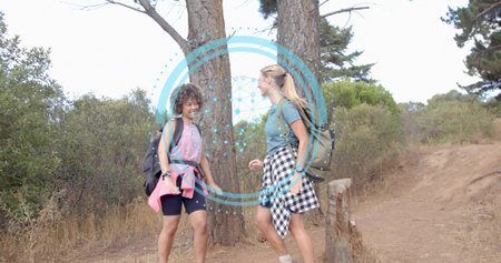 Walking two women talking on dirt trail, carrying backpacks, tied shirts, pine trunks, blue overlay. Hikers, hiking, forest, sunlit, outdoors, nature, packsの写真素材