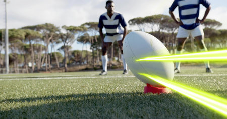 Balancing white rugby ball on red tee on grass pitch, neon streaks, players wearing striped jerseys. Field, athletes, outdoors, teamwork, focus, preparation, goalpostsの写真素材