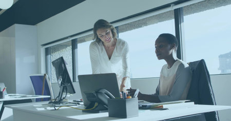Collaborating two women in business attire leaning over laptop at desk near windows, with monitor. Teamwork, colleagues, workstation, daylight, modern, urban, corporateの写真素材