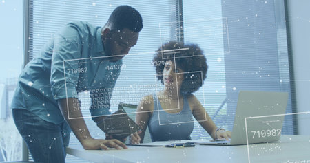 Leaning man holding tablet, seated woman sleeveless gray writing at office desk, laptop AR overlays. Colleagues, collaboration, analysts, data, graphics, daylight, blindsの写真素材