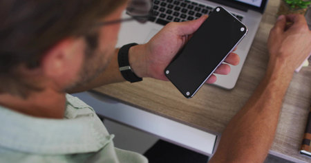 Holding smartphone, man scrolling at wooden desk with laptop, wearing green shirt and wristwatch....