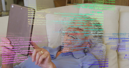 Reclining senior woman tapping tablet on home sofa, wearing light blue collared shirt, showing code. Elderly, holographic, coding, relaxed, cozy, lounge, cushionsの写真素材
