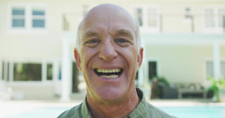 Smiling senior man wearing light green collared shirt posing poolside by two-story house balcony....