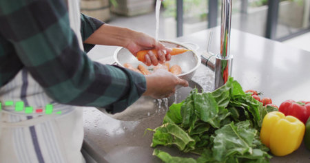 Washing woman wearing striped apron rinsing carrots in colander under chrome faucet at kitchen sink. Produce, vegetables, daylight, natural, modern, stainless, countertopの写真素材