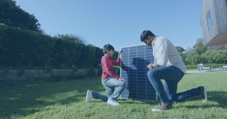 Kneeling mature man and boy inspecting vertical solar panel on backyard lawn, showing wristwatch....