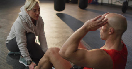 Performing situps man in red top on rubber floor, trainer in hoodie holding ankles with smartwatch. Gym, crunches, athlete, coach, weights, focus, lightingの写真素材