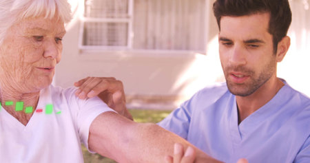 Supporting nurse in blue scrubs holding senior woman in white top extending right arm at home. Caregiver, elderly, outdoors, residential, assistance, movement, therapyの写真素材