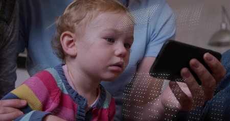 Watching infant girl gazing at smartphone at home, in striped sweater, supported by adult hands. Baby, family, parents, device, screen, cozy, softの写真素材