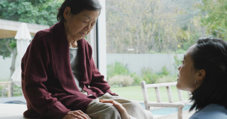 Comforting adult woman touching senior woman's knee by glass door, burgundy cardigan, copy space. Caregiving, tenderness, comfort, calm, home, patio, gardenの写真素材