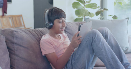 Sitting boy wearing pink t-shirt and jeans holding tablet on living room sofa, with gray headphones....