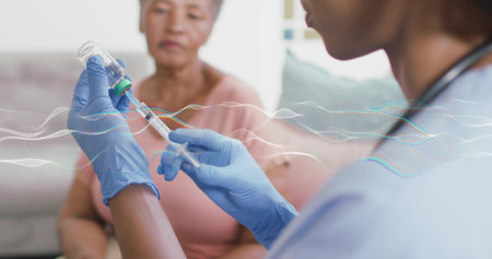 Preparing nurse in blue scrubs and gloves drawing vaccine into syringe at home with stethoscope. Caregiver, clinician, patient, elderly, livingroom, medical, glassの写真素材