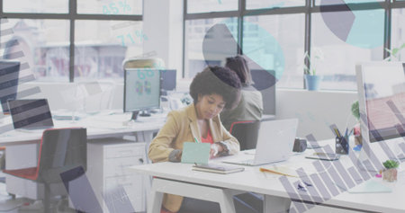 Writing woman with curly hair wearing tan blazer working at open-plan desk, with laptop and charts....