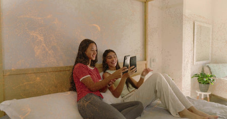 Smiling, two women scrolling smartphone on bed with wood headboard, wearing red tee and white tank....