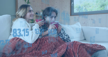 Smiling women in sweatshirt plaid cuddling under rust blanket sipping red mugs at home. Cozy,...
