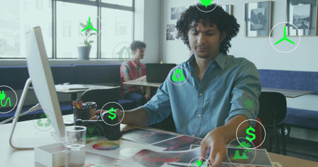 Reviewing curly-haired designer in blue shirt arranging printed proofs and color wheel, green icons. Coworking, studio, printouts, palette, laptop, daylight, plantの写真素材