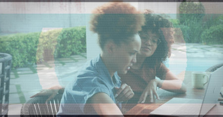 Leaning two midadult women in short-sleeve shirt and glasses, working over laptop and mug on patio....