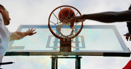 Reaching duo in white-red tops dueling for rebound at park under backboard, ball through rim. Hoop,...