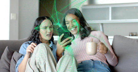 Sitting two women sharing smartphone on couch in living room, ceramic mug and green AR particles....