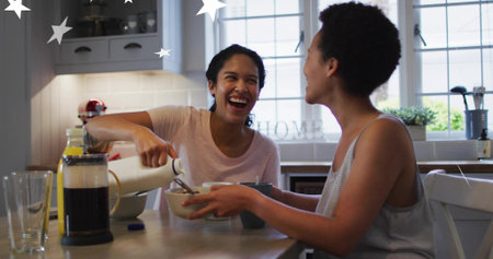 Pouring from white pitcher, women laughing at breakfast at kitchen table in pink and striped tops. Duo, friendship, morning, daylight, cozy, coffee, candidの写真素材