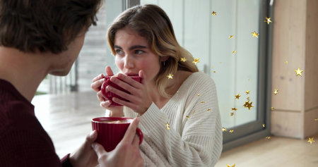 Sipping woman in cream knit sweater holding red mug near glass doors, with gold stars. Couple,...