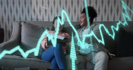 Smiling couple on gray sofa at home, woman in sweater, jeans leaning toward tablet, chart overlay....