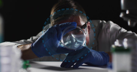 Leaning male lab worker wearing full PPE inspecting petri dish at bench, with blue scanning overlay. Specimen, inspection, precision, research, science, microscope, vialの写真素材