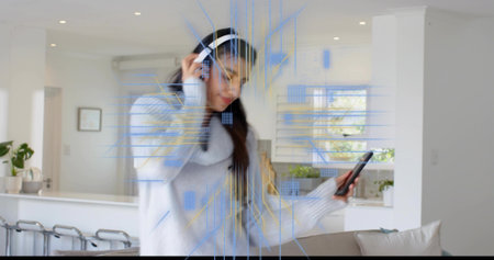 Listening woman in gray-sweater touching white headset in kitchen, holding phone with AR overlay....