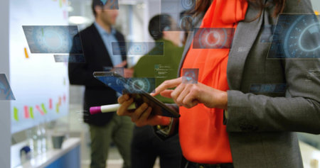 Using tablet, woman wearing grey blazer, orange blouse pointing with pink-marker in office, HUD....