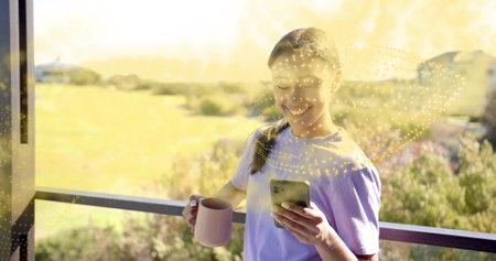 Smiling woman holding mug and checking smartphone on balcony wearing purple T-shirt, copy space....