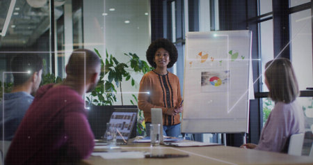 Presenting woman in rust blouse and jeans standing at flip chart in meeting room with laptops....