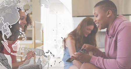 Showing rightmost woman wearing mauve hoodie holding smartphone and cards at home, globe overlay. Friends, phone, map, worldmap, casual, cozy, daylightの写真素材