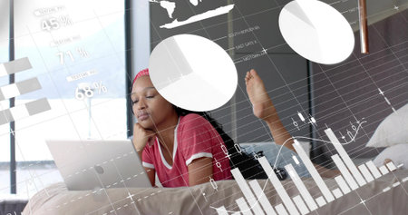 Lying woman in red T-shirt and pink headband using laptop in bedroom with earbuds, data overlays. Focus, study, remote, charts, graphs, daylight, deviceの写真素材